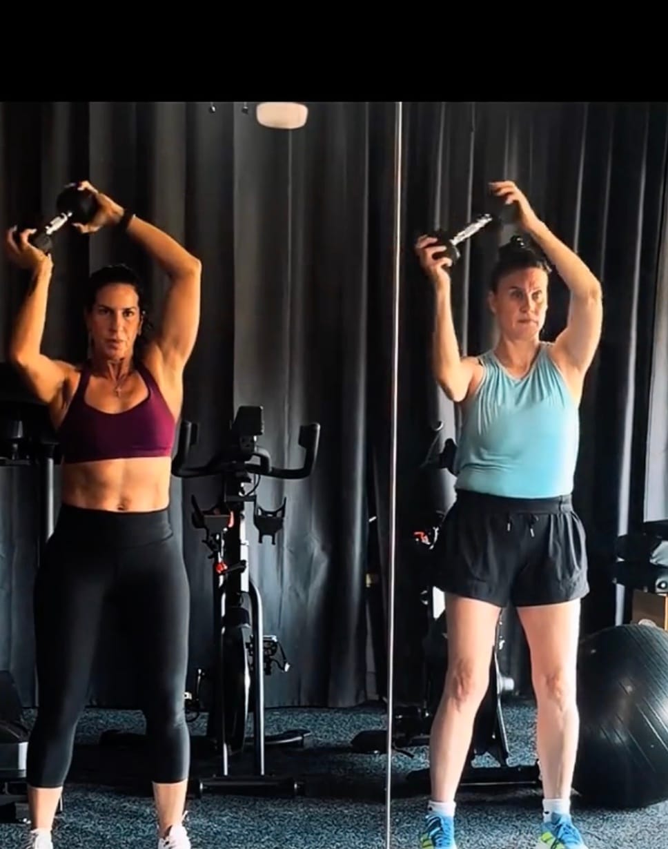 Coach Lala and a client side by side, both holding dumbbells overhead in matching strength postures.